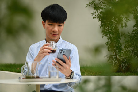 Portrait of Young Asian man using phone while drinking a coffee and sitting at the outdoor.