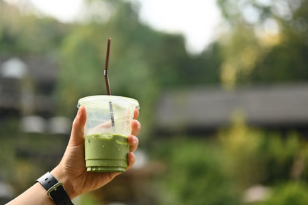 Close-up image of a male hand holding a Matcha latte or Green tea latte in a plastic glass with a scene of a cafe in nature.の写真素材