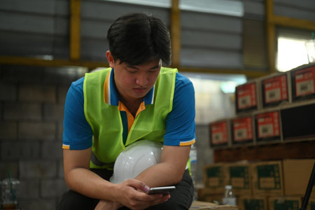 A warehouse worker male on break relaxes and reads the news on his smart phone in an industrial...