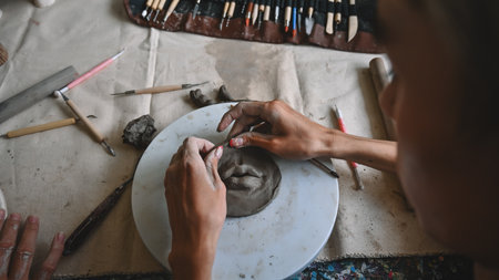 An overhead view of hands shaping a clay piece on a pottery wheel, surrounded by various sculpting tools and clay pieces, Crafts and DIY with people concept.の写真素材