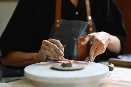 A person working on a pottery wheel, shaping clay with both hands and using a sculpting tool. They wear an apron, focusing intently on the clay piece in progress, Crafts and DIY with people concept.の写真素材