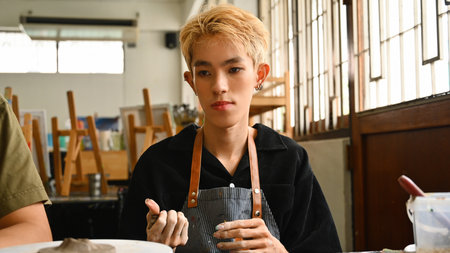 A young LGBT artist with blonde hair focuses on shaping clay in a bright studio. The background includes art supplies and easels, indicating a creative environment, Crafts and DIY with people concept.の写真素材