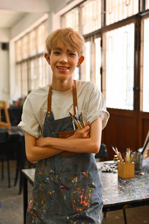 A smiling Young LGBT artist, wearing a paint-splattered striped apron and holding paintbrushes, stands confidently in a brightly lit art studio with tables, art supplies, and large windows in the background.の写真素材