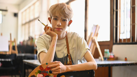 A confident LGBT artist with blonde hair, wearing a paint-splattered striped apron, sits holding a...