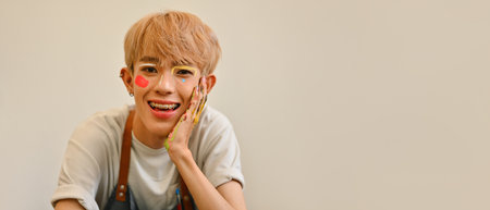 Young LGBT artist boy with a painted face and hands, smiling brightly and Happily in a white shirt and apron, Creativity and joy with copy space, showcasing a colorful and playful approach to art.の写真素材