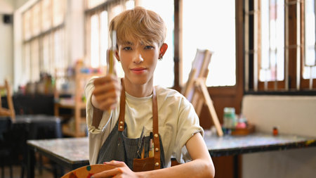 A confident LGBT artist holds up a paintbrush towards the camera, aims, and estimates in a spacious art studio filled with light and supplies.の写真素材