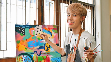 A joyful LGBT artist boy with blonde hair, wearing a paint-splattered striped apron, smiles while holding paintbrushes and working on a vibrant abstract painting in a bright, creative art studio.の写真素材