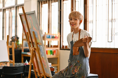 A cheerful LGBT artist boy with blonde hair, wearing a striped apron, sits in front of an easel, holding a palette in a bright, spacious art studio filled with light and creative energy.の写真素材