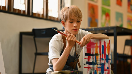 A focused LGBT artist with blonde hair, wearing a striped apron, sits on the floor painting a colorful abstract canvas in a bright, spacious art studio filled with light and creative energy.の写真素材