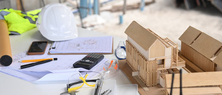 A construction site table with architectural models, blueprints, a hard hat, a calculator, safety...