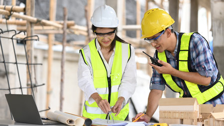 An engineer uses a compass on blueprints while meeting with a foreman at a construction site, Discuss project details, and are surrounded by tools, a laptop, and a scale model.の写真素材