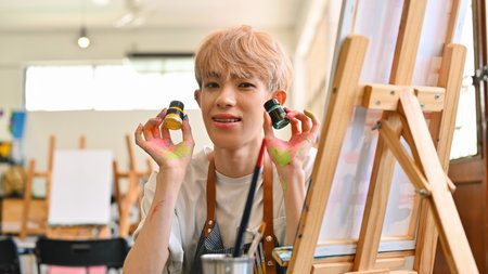 A young LGBT artist in a studio proudly displays yellow and green paint bottles with a cheerful smile. Their hands are covered in colorful paint, reflecting their joyful and creative process..の写真素材