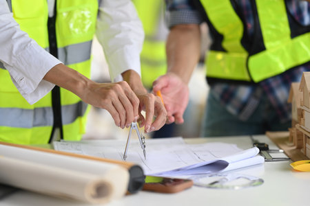 Close-up of a hand using a divider compass on blueprints at a construction site. The Two professional construction workers focus on precise measurements and planning, surrounded by tools and documents.の写真素材