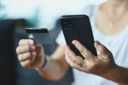 Close-up mature woman using a phone, holding a credit or debit card, making internet payments,...