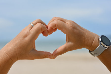 Cropped image of mature adult hands making heart shape over the beach and sea on the background.の写真素材