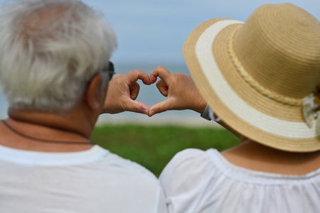 Rear view with Elderly Asian couple making head shapes with their hands together in front of the beach.の写真素材
