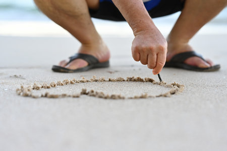 Close-up man's hand drawing heart shape on sea sand.の写真素材