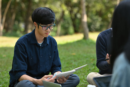 A focused university student reviewing learning lessons in the textbook while sitting with his...