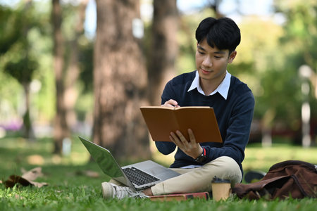 A focused university student taking a note while reviewing learning lessons on the laptop and...