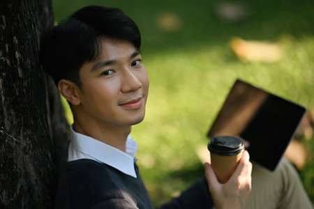 Close-up image with Young Asian university student man sitting under the tree while holding a coffee and tablet.の写真素材