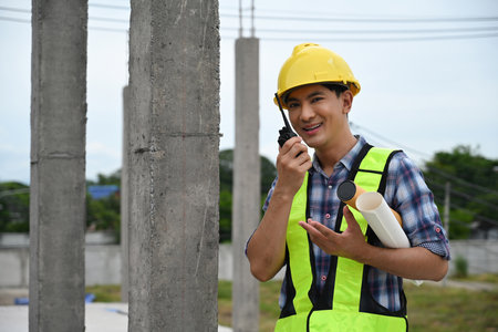 A Foreman using a walkie-talkie on a construction site, holding blueprints, coordinating and...