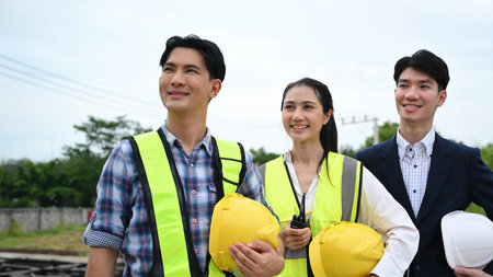 Asian Construction worker team standing in a row, smiling confidently on-site, holding helmets and...