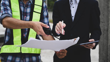 Close-up of a foreman and manager reviewing construction plans on-site, using a walkie-talkie and...