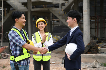 A contractor and a businessman shake hands at a construction site, with a female worker clapping in...