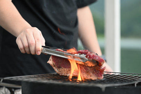 Searing Steak Over an Open Flame, A hand using tongs to hold a seasoned steak over a grill with visible flames, capturing the intense heat and sizzle of outdoor grilling.の写真素材