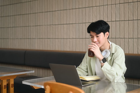 A Young student enjoys a coffee while learning with a laptop in the comfort of studying from their...