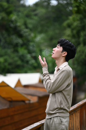 A young man breathes cold air as he looks into the distance amidst greenery, enjoying a quiet moment...