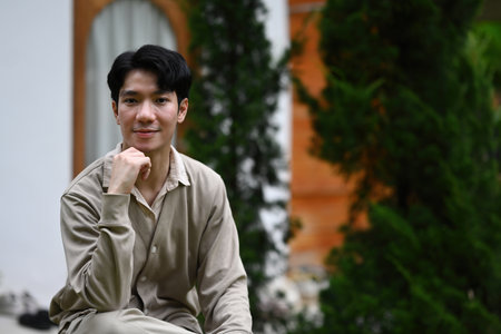 A Portrait of a Young Man sits outdoors, resting his chin on his hand while wearing a soft...