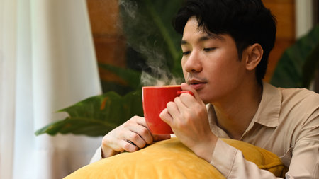 A Young man leans into his steaming red mug, inhaling the warm scent while nestled comfortably on a...