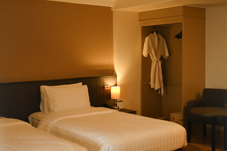 A cozy hotel room featuring twin beds, warm lighting, a bathrobe hanging in the wardrobe, and a small lounge chair with a table, creating a restful atmosphere.の写真素材