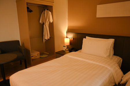 A warm and cozy hotel room featuring a comfortable bed, bedside lamp, open wardrobe with a bathrobe, and a seating area for relaxation, designed.の写真素材