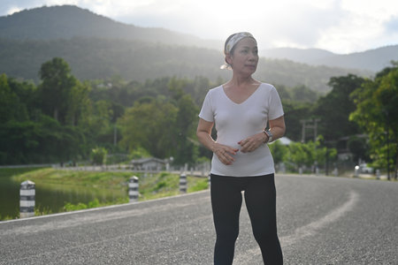 An old woman stands on a peaceful road with scenic mountain views, taking a moment to pause and...