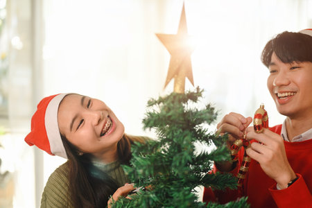 A happy couple in Santa hats decorates a Christmas tree, sharing laughter and joy as they add...