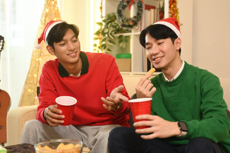 Two friends, wearing festive hats, enjoy a lighthearted conversation while sipping drinks and...