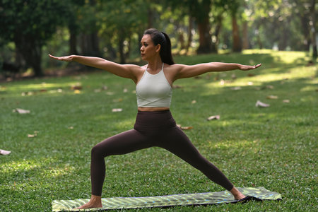 An Asian woman demonstrates Virabhadrasana II, extending her arms and gazing forward with confidence, embodying strength, balance, and focus. Yoga workout in a peaceful outdoor.の写真素材