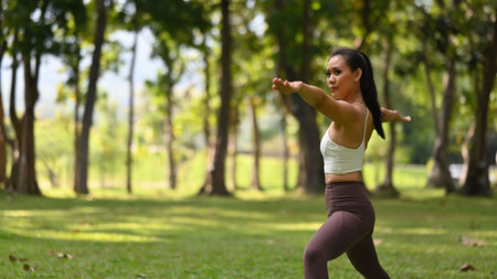 A woman performs Warrior II pose outdoors, arms extended parallel to the ground, front knee bent, and gaze over the front hand, exuding focus and balance amidst nature.の写真素材