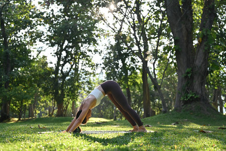An Asian sporty woman practices the Downward Facing Dog yoga pose in a park, promoting flexibility,...