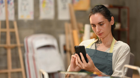 An artist in an apron focuses on her smartphone while seated in a creative studio filled with...