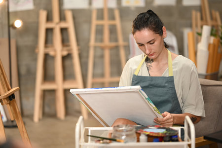 A Russian female artist sits focused on her canvas, with a paintbrush in hand, surrounded by...