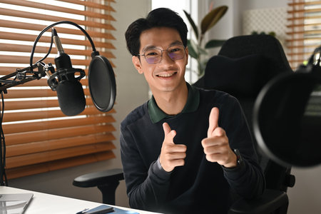 A smiling podcast host in a modern home studio gives a double thumbs up while sitting in front of a...