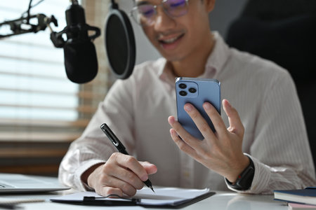 A professional man in a home studio writes notes while checking his smartphone, surrounded by a microphone, laptop, and office essentials, showcasing productivity and remote work.の写真素材
