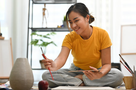 An Asian woman in a yellow shirt sits on the floor, holding a palette and brush, carefully observing a vase and an apple while preparing to paint, surrounded by art supplies in a bright room.の写真素材