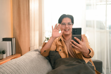 A man sitting on a couch smiles and waves while video calling on his smartphone, reflecting modern communication, remote connection, and digital interaction in a cozy home setting.の写真素材