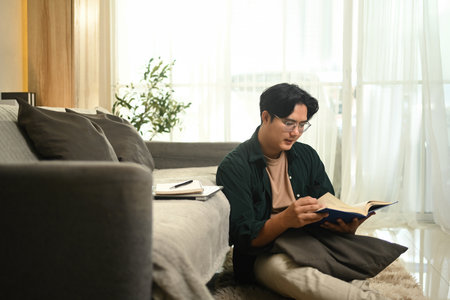 A young man sits comfortably on the floor, relaxing with a favorite book, reading a book in a cozy...