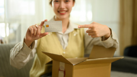 A Close-up of a happy woman holds up her credit card, pointing at it while unboxing her online...