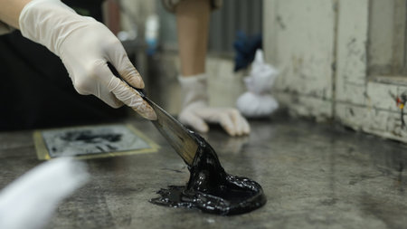 A close-up of an artist's hands spreading thick black ink on a workbench, Preparing Ink for...